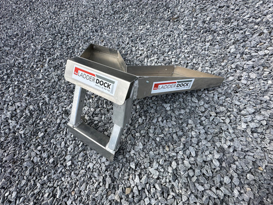 A silver-colored metal corner dock designed for ladder attachment, placed on a gravel surface.
