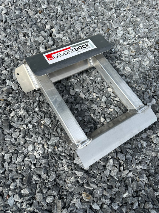 A silver-colored ladder rung attachment lying on a gravel surface, with the brand name 'Ladder Dock' visible on it.